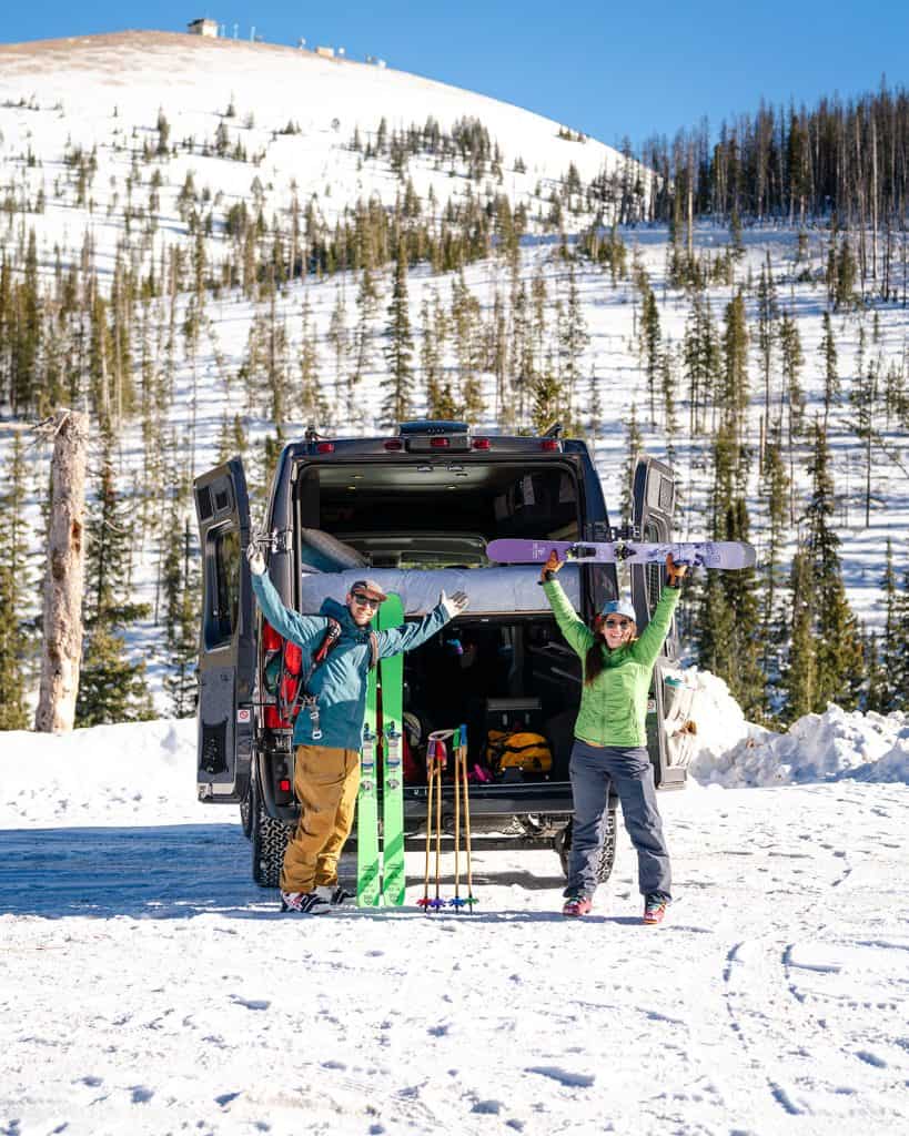 A couple standing at the back of their camper van with ski gear on excited to hit the slopes.