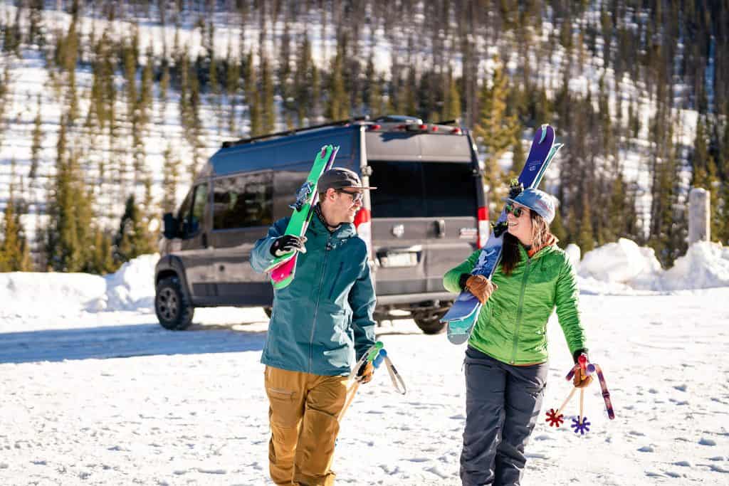 Man and woman walking away from their camper van at a ski resort with ski gear in hand and smiling at one another.